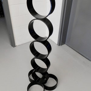 Puede incluir: Una escultura moderna compuesta por anillos negros brillantes apilados. Los anillos est&aacute;n dispuestos verticalmente, creando una estructura similar a una torre. La escultura se encuentra sobre un fondo neutro, resaltando su forma y dise&ntilde;o.