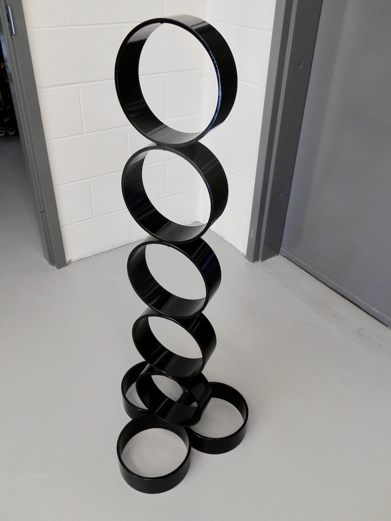 Puede incluir: Una escultura moderna compuesta por anillos negros brillantes apilados. Los anillos est&aacute;n dispuestos verticalmente, creando una estructura similar a una torre. La escultura se encuentra sobre un fondo neutro, resaltando su forma y dise&ntilde;o.