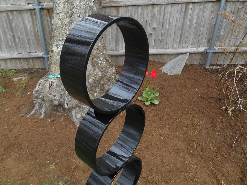 Puede incluir: Una escultura exterior negra y moderna compuesta por tres formas circulares brillantes e interconectadas. La escultura se encuentra verticalmente en un jard&iacute;n, con un tronco de &aacute;rbol y una valla de madera en el fondo. Los tulipanes rojos florecen cerca.