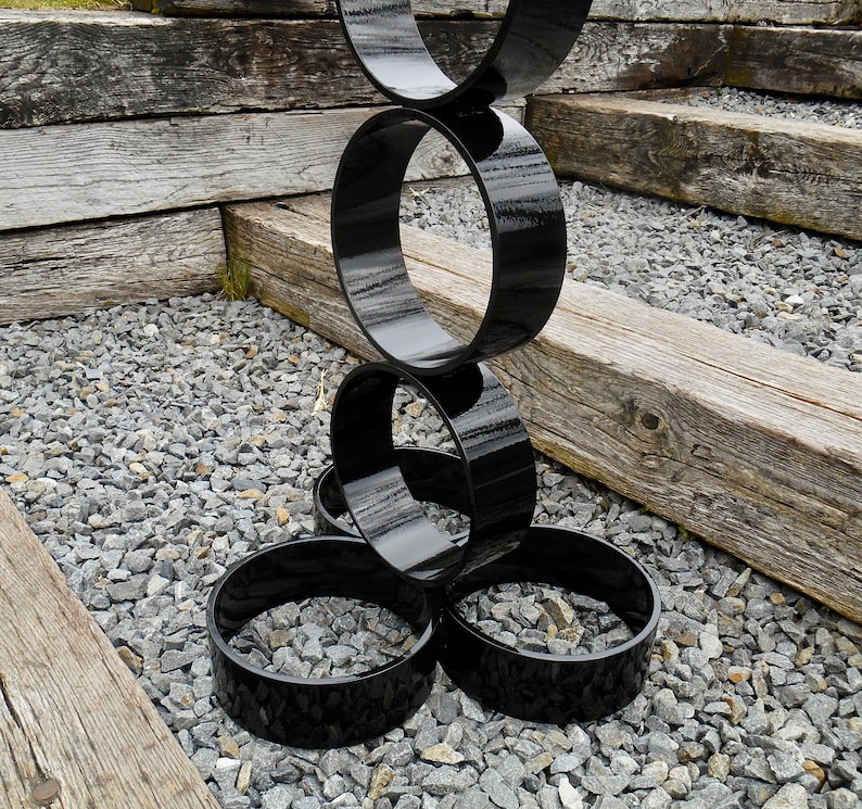Puede incluir: Una escultura moderna para exteriores compuesta por anillos negros brillantes interconectados. La escultura se encuentra sobre un lecho de peque&ntilde;as piedras grises, con vigas de madera envejecida en el fondo, creando un contraste de texturas y colores.