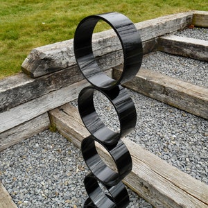 Puede incluir: Una escultura moderna para exteriores compuesta por cuatro anillos negros brillantes apilados. La escultura se encuentra sobre vigas de madera desgastadas, peque&ntilde;as piedras grises y hierba verde, creando un contraste de texturas y colores.