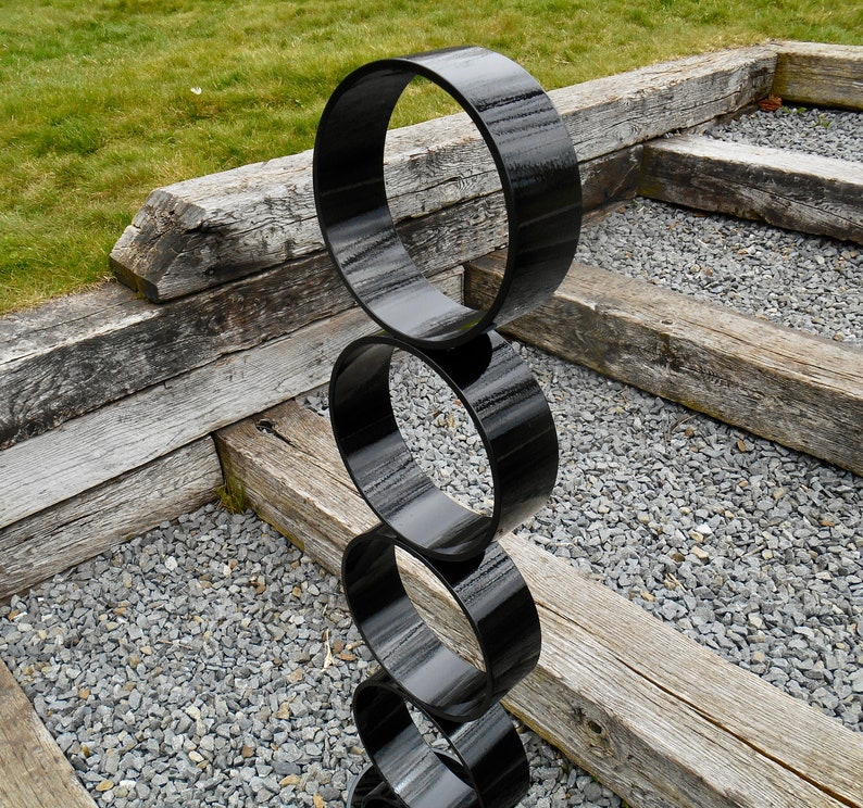 Puede incluir: Una escultura moderna para exteriores compuesta por cuatro anillos negros brillantes apilados. La escultura se encuentra sobre vigas de madera desgastadas, peque&ntilde;as piedras grises y hierba verde, creando un contraste de texturas y colores.