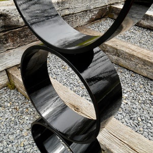 Puede incluir: Una escultura moderna para exteriores compuesta por tres formas circulares negras brillantes e interconectadas. La escultura se encuentra sobre un fondo de vigas de madera envejecida y peque&ntilde;as piedras grises, creando un contraste de texturas.