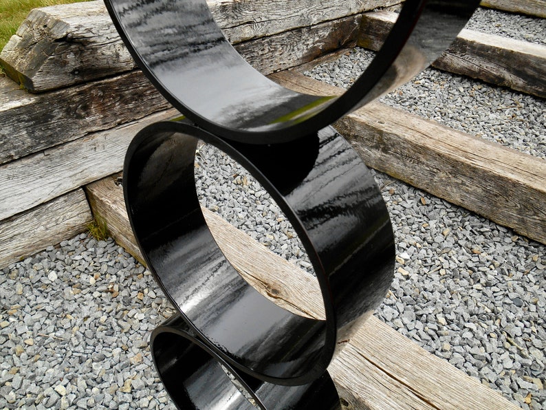 Puede incluir: Una escultura moderna para exteriores compuesta por tres formas circulares negras brillantes e interconectadas. La escultura se encuentra sobre un fondo de vigas de madera envejecida y peque&ntilde;as piedras grises, creando un contraste de texturas.