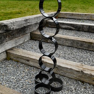 Puede incluir: Una escultura exterior negra y moderna compuesta por anillos circulares brillantes e interconectados. La escultura se alza sobre un lecho de grava gris, con escalones de madera desgastada y un c&eacute;sped verde al fondo. Los anillos var&iacute;an en tama&ntilde;o.
