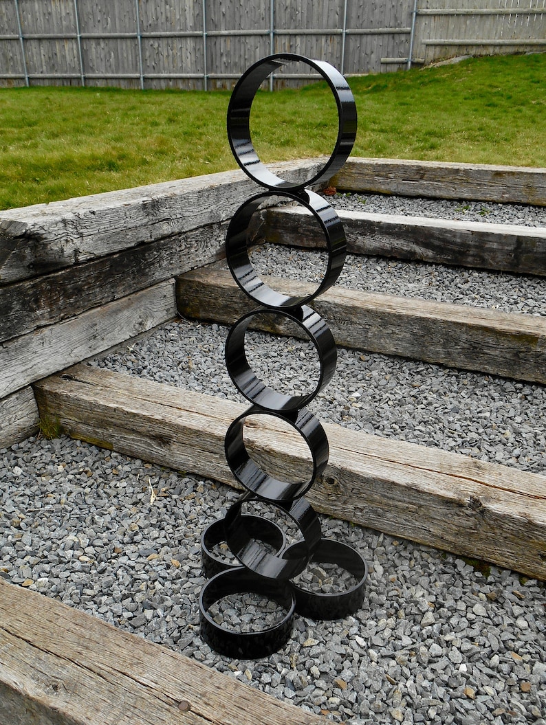 Puede incluir: Una escultura exterior negra y moderna compuesta por anillos circulares brillantes e interconectados. La escultura se alza sobre un lecho de grava gris, con escalones de madera desgastada y un c&eacute;sped verde al fondo. Los anillos var&iacute;an en tama&ntilde;o.