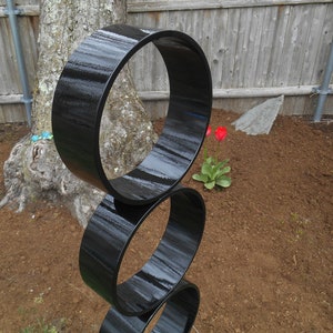 Puede incluir: Escultura moderna para exteriores compuesta por tres anillos negros brillantes apilados. La escultura est&aacute; ubicada en un parterre con un tronco de &aacute;rbol y una valla de madera al fondo. Los tulipanes rojos a&ntilde;aden un toque de color.