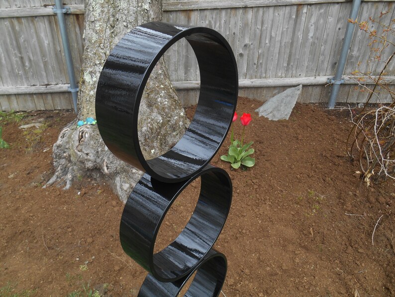 Puede incluir: Escultura moderna para exteriores compuesta por tres anillos negros brillantes apilados. La escultura est&aacute; ubicada en un parterre con un tronco de &aacute;rbol y una valla de madera al fondo. Los tulipanes rojos a&ntilde;aden un toque de color.