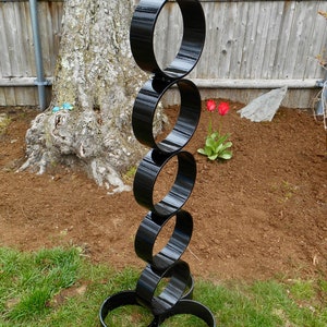 Puede incluir: Una escultura alta de metal negro compuesta por anillos interconectados. La escultura se encuentra sobre un c&eacute;sped, con un &aacute;rbol y una valla de madera al fondo. Tambi&eacute;n se ven dos tulipanes rojos.