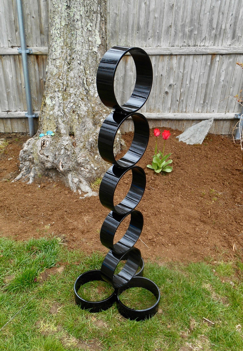 Puede incluir: Una escultura alta de metal negro compuesta por anillos interconectados. La escultura se encuentra sobre un c&eacute;sped, con un &aacute;rbol y una valla de madera al fondo. Tambi&eacute;n se ven dos tulipanes rojos.