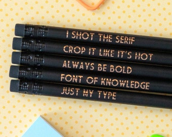 May include: Five black pencils with gold lettering. The pencils have the following text: "I shot the serif", "Crop it like it's hot", "Always be bold", "Font of knowledge", and "Just my type".