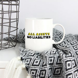 May include: White ceramic mug with black text that reads "ALL ASSETS NO LIABILITIES" in yellow outlined letters. The mug is sitting on a black and white patterned blanket.