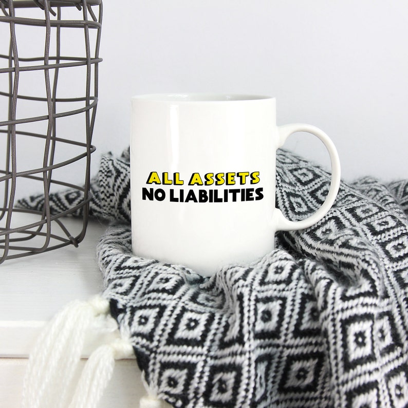 May include: White ceramic mug with black text that reads "ALL ASSETS NO LIABILITIES" in yellow outlined letters. The mug is sitting on a black and white patterned blanket.