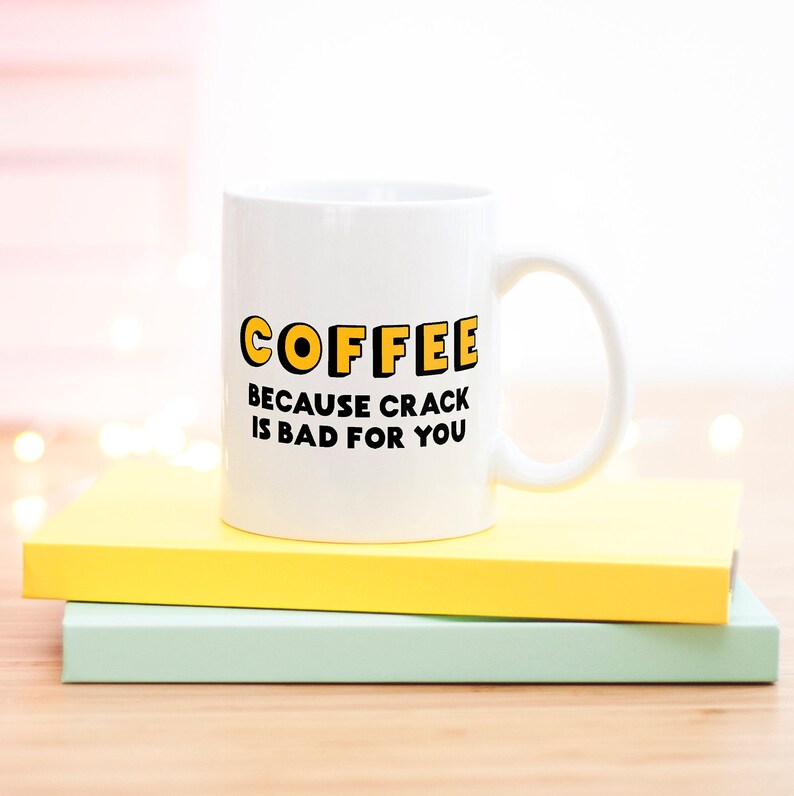 K&ouml;nnte beinhalten: Wei&szlig;e Kaffeetasse mit schwarzem Text, der "COFFEE BECAUSE CRACK IS BAD FOR YOU" in gelb umrandeten Buchstaben lautet.