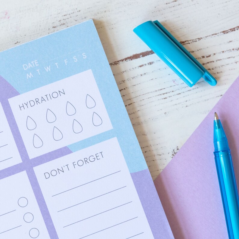 Op de afbeelding: Een blauwe en paarse planner met een hydratatietracker en een "Niet vergeten"-sectie. De planner heeft een blauwe pen en een blauwe markeerstift erop.