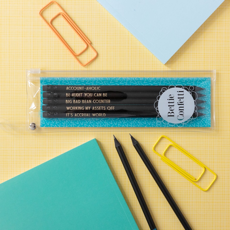 May include: A clear plastic pencil case with a blue background and white stars. The case contains five black pencils with gold lettering that reads "ACCOUNT-AHOLIC", "BE AUDIT YOU CAN BE", "BIG BAD BEAN COUNTER", "WORKING MY ASSETS OFF", and "IT'S ACCRUAL WORLD". The case also has a round sticker with the text "Bettie Confetti" in black and white.