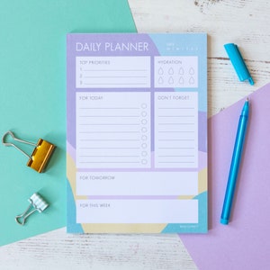 Op de afbeelding: Een dagelijkse planner met een geometrisch ontwerp in paars, blauw en geel. De planner heeft secties voor topprioriteiten, hydratatie, voor vandaag, niet vergeten, voor morgen en voor deze week. De planner ligt op een wit houten oppervlak met een blauwe pen en twee gouden paperclips.