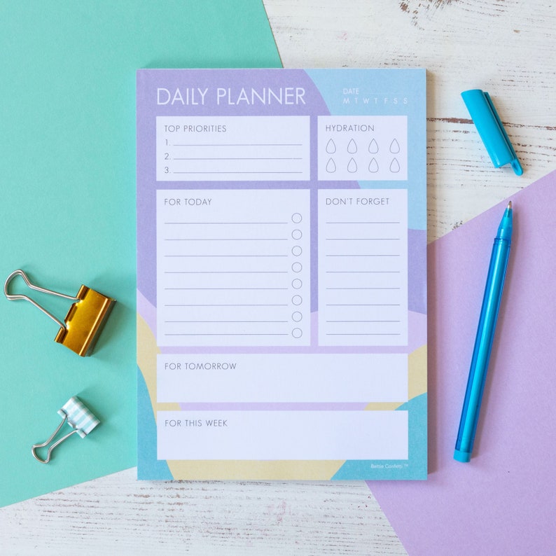 Op de afbeelding: Een dagelijkse planner met een geometrisch ontwerp in paars, blauw en geel. De planner heeft secties voor topprioriteiten, hydratatie, voor vandaag, niet vergeten, voor morgen en voor deze week. De planner ligt op een wit houten oppervlak met een blauwe pen en twee gouden paperclips.