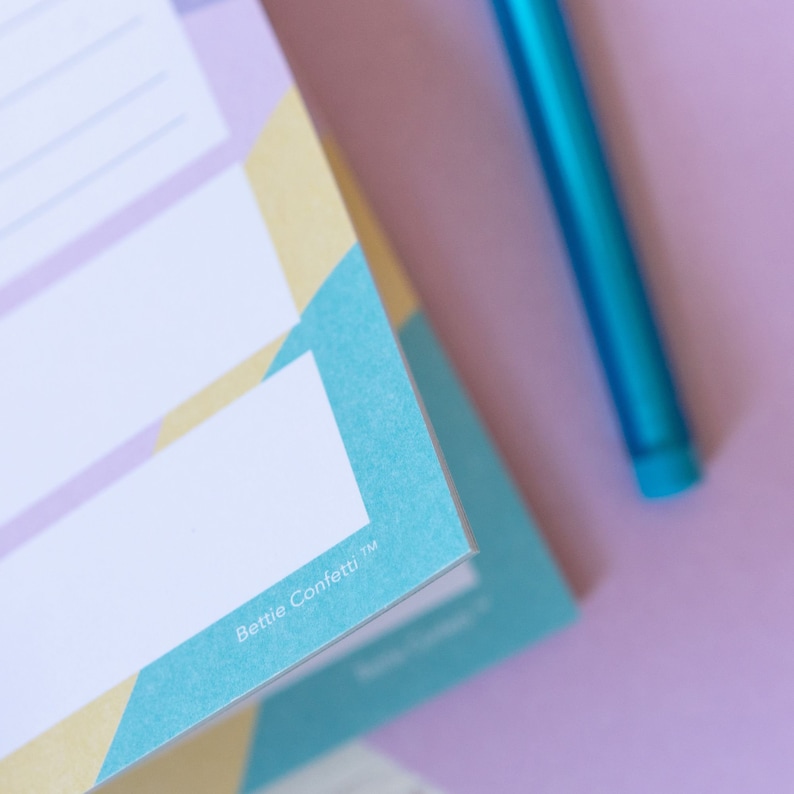 Op de afbeelding: Een close-up van een blauwe en gele planner met de tekst "Bettie Confetti&trade;" op de omslag. De planner is open op een pagina met lijnen om te schrijven.