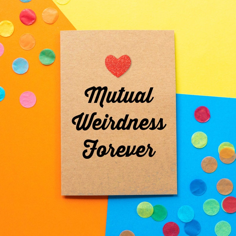 May include: A brown card with the text "Mutual Weirdness Forever" in black cursive font. A red glitter heart is above the text. The card is on a background of yellow, orange, and blue paper with confetti.