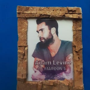 Può includere: Un'immagine incorniciata con Adam Levine dei Maroon 5. L'immagine è inserita in una cornice rettangolare realizzata in un materiale marrone chiaro strutturato. Il testo "Adam Levine" e "Maroon 5" è visibile sull'immagine.