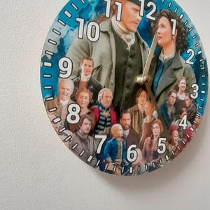 Può includere: Orologio da parete rotondo con bordo blu e numeri bianchi, con un collage di personaggi. Le lancette segnano le 12. L'orologio è appeso a una parete bianca.