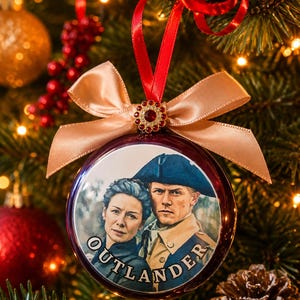 May include: A round, dark red Christmas bauble featuring a printed image of a woman and a man in historical attire with the text OUTLANDER. The bauble is topped with a champagne-coloured satin bow and a small, circular red rhinestone accent.