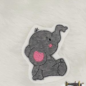 Könnte beinhalten: Eine graue gestickte Elefanten-Applikation mit einem rosa Herz auf dem Bauch. Der Elefant sitzt mit erhobenem Rüssel.