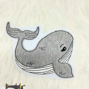 Könnte beinhalten: Eine graue Wal-Applikation auf weißem Hintergrund. Der Wal hat ein lächelndes Gesicht und eine detaillierte Schwanzflosse.