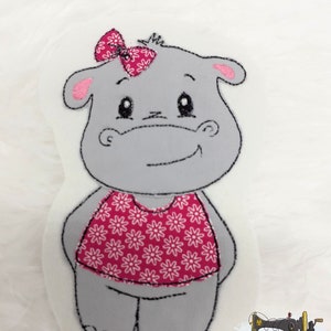 Könnte beinhalten: Ein graues Plüsch-Nilpferd-Spielzeug mit einer rosa Schleife und einem rosa Oberteil mit Blumenmuster. Das Nilpferd hat schwarze, gestickte Umrisse und einen lächelnden Ausdruck. Das Spielzeug befindet sich auf weißem Hintergrund. Das Bild enthält auch ein Logo mit Text.