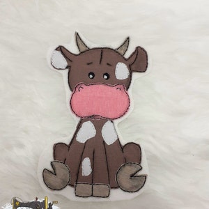 Könnte beinhalten: Eine braune und weiße Kuh-Applikation mit rosa Nase und weißen Flecken. Die Kuh sitzt mit gekreuzten Beinen und hat einen freundlichen Ausdruck.