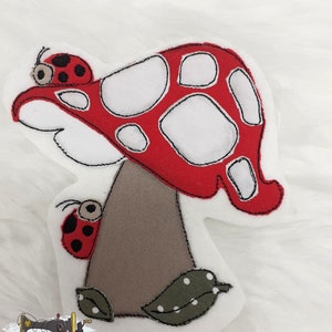 Könnte beinhalten: Ein skurriler Plüschpilz mit rotem Hut und weißen Punkten sowie einem braunen Stiel. Zwei Marienkäfer mit schwarzen Punkten und ein grünes Blatt mit weißen Punkten sind ebenfalls vorhanden. Auf weißem Hintergrund.