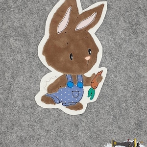 Könnte beinhalten: Ein braunes Filzkaninchen mit blauer Latzhose und weißen Punkten, das eine Karotte hält. Das Kaninchen steht auf einem grauen Filzgrund.