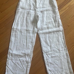 Puede incluir: Pantalones de lino blancos con diseño de pernera ancha. Los pantalones tienen cierre de botón y cremallera, trabillas para el cinturón y bolsillos. La tela parece ligera y transpirable, adecuada para climas cálidos.