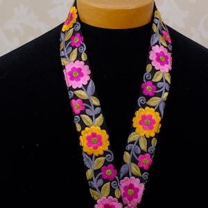 Pode incluir: Fita preta com flores bordadas em tons de rosa, amarelo e verde. As flores são dispostas em um padrão repetitivo ao longo da fita.