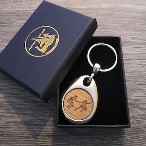 Op de afbeelding: Een zilveren ovale sleutelhanger met een houten inlay met een silhouet van twee dansende mensen. De sleutelhanger zit in een zwarte doos met een gouden logo met de tekst "Jorkvik Wood Handmade in York".