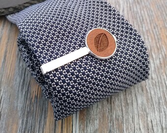May include: A silver tie clip with a wooden leaf design. The wooden piece is round and has a natural wood grain.
