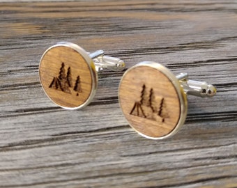 May include: A pair of wooden cufflinks with a silver metal backing. The cufflinks feature a design of a tent and trees.