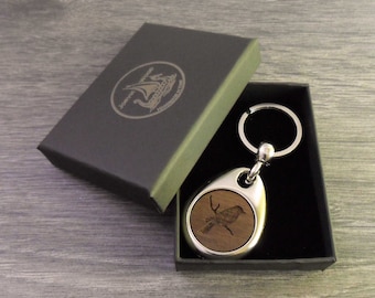 Puede incluir: Un llavero de plata y madera con diseño de pájaro, presentado en una caja de regalo gris oscuro. El llavero presenta una incrustación circular de madera con un grabado detallado de un pájaro. La caja tiene el texto "JORVIK WOOD" y un logotipo.