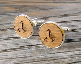 May include: A pair of round cufflinks with a silver border and a wooden centre. Each wooden centre features an engraved image of a goose and a gosling. The cufflinks are displayed on a wooden surface.
