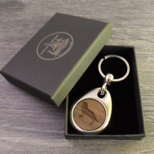 Puede incluir: Un llavero de metal plateado y madera con la silueta de un perro dentro de un colgante en forma de lágrima. El llavero se presenta en una caja gris oscuro con el texto "Jorvik Wood Handmade Gifts" y un logotipo de barco vikingo.