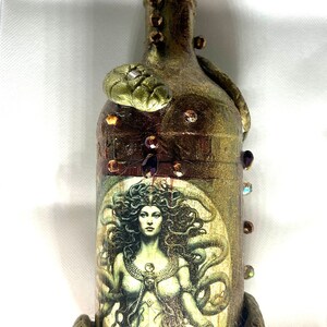 May include: A decorative glass bottle with a gold and brown finish, featuring a printed image of Medusa, the Greek mythological figure with snakes for hair. The bottle is adorned with gold and green snakes, and embellished with rhinestones.
