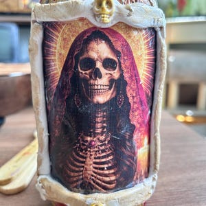 May include: A decorative bottle featuring a skull image with a red veil and a skeleton. The bottle has a gold-colored top and is adorned with small skull-shaped embellishments. The image is framed with a cream-colored border.