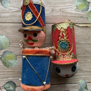 May include: Three vintage Christmas ornaments shaped like toy soldiers. The ornaments feature blue, red, and gold accents, with painted faces and decorative hats. Each ornament has a gold string for hanging.