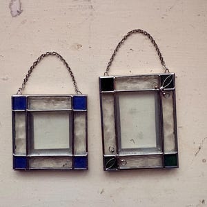 May include: Two small, square stained glass frames with silver chain hangers. One frame has blue accents, the other has green accents. Both have clear glass centers and are set against a light-colored background.