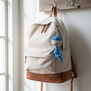 Op de afbeelding: Een beige rugzak met bruine leren details en riemen hangt aan een houten haak. Een blauw-witte gehaakte sleutelhanger in de vorm van een lachend wezen is bevestigd aan de rits van de rugzak.