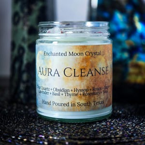 May include: A glass jar candle with a label that reads "Aura Cleanse." The label has a watercolor design with the words "Enchanted Moon Crystal." Ingredients listed include quartz, obsidian, and herbs. The candle is hand-poured in South Texas.