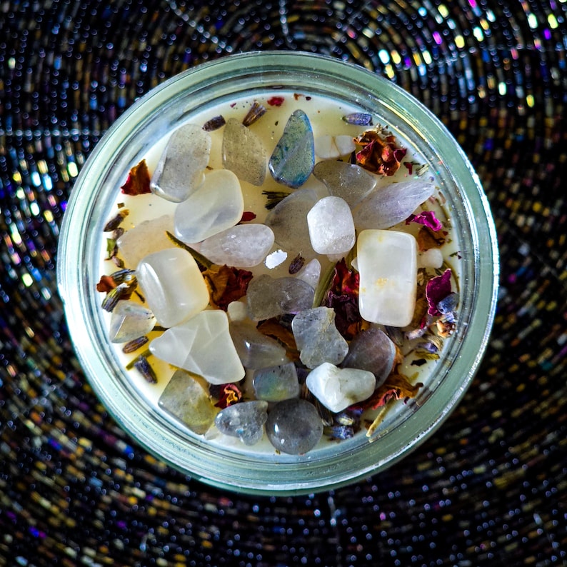 Op de afbeelding: Een ronde glazen potkaars gevuld met een romige wasbasis, versierd met diverse gepolijste edelstenen in tinten wit, grijs en blauw. Gedroogde bloemen en kruiden zijn verspreid. De kaars staat op een donkere, getextureerde ondergrond.