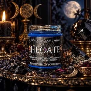 Hand-poured Hecate goddess candle photographed in a mystical gothic setting. The cobalt blue glass candle jar features an elaborate black and antique gold celestial label, surrounded by black crystals, lavender, dried botanicals, glowing candlelight, and a luminous full moon in the background. Inspired by Hecate, the Greek goddess of witchcraft and crossroads, this crystal intention candle evokes moon magic, shadow work, divination, spiritual protection, and transformation