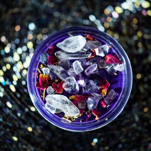Pu&ograve; includere: Una candela viola in un contenitore di vetro trasparente, riempita con cristalli chiari e ametista e petali di rosa secchi. Lo sfondo &egrave; scuro e scintillante. La candela ha un bordo viola.